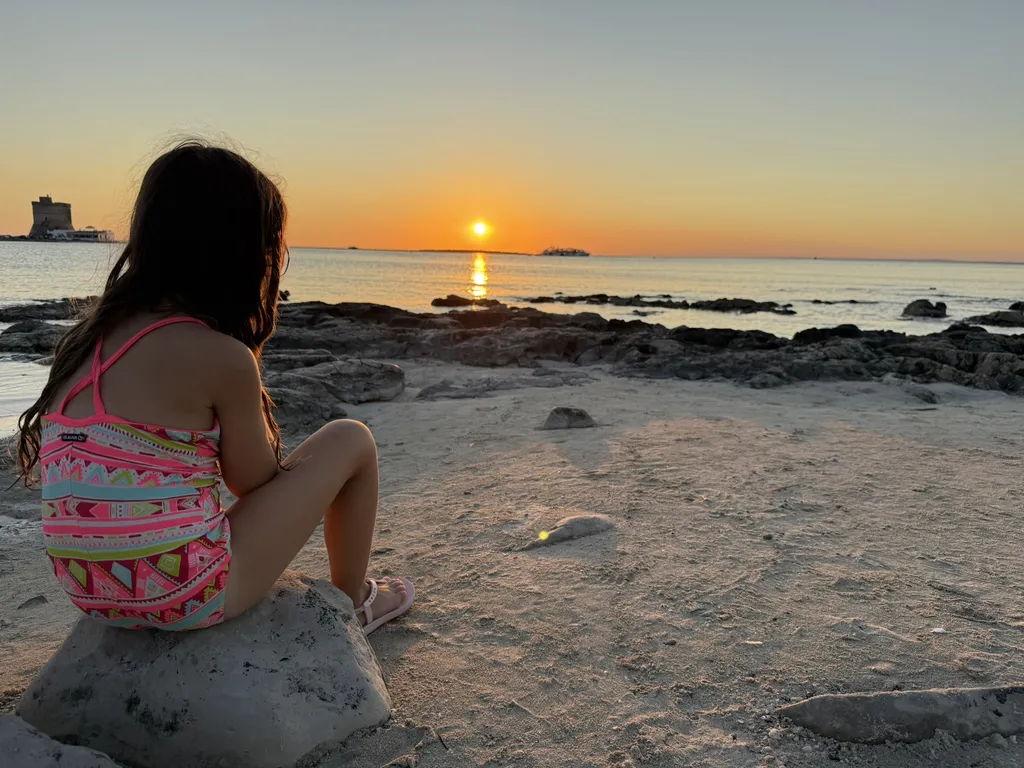 Un bambino seduto su uno scoglio guarda il tramonto sul mare.