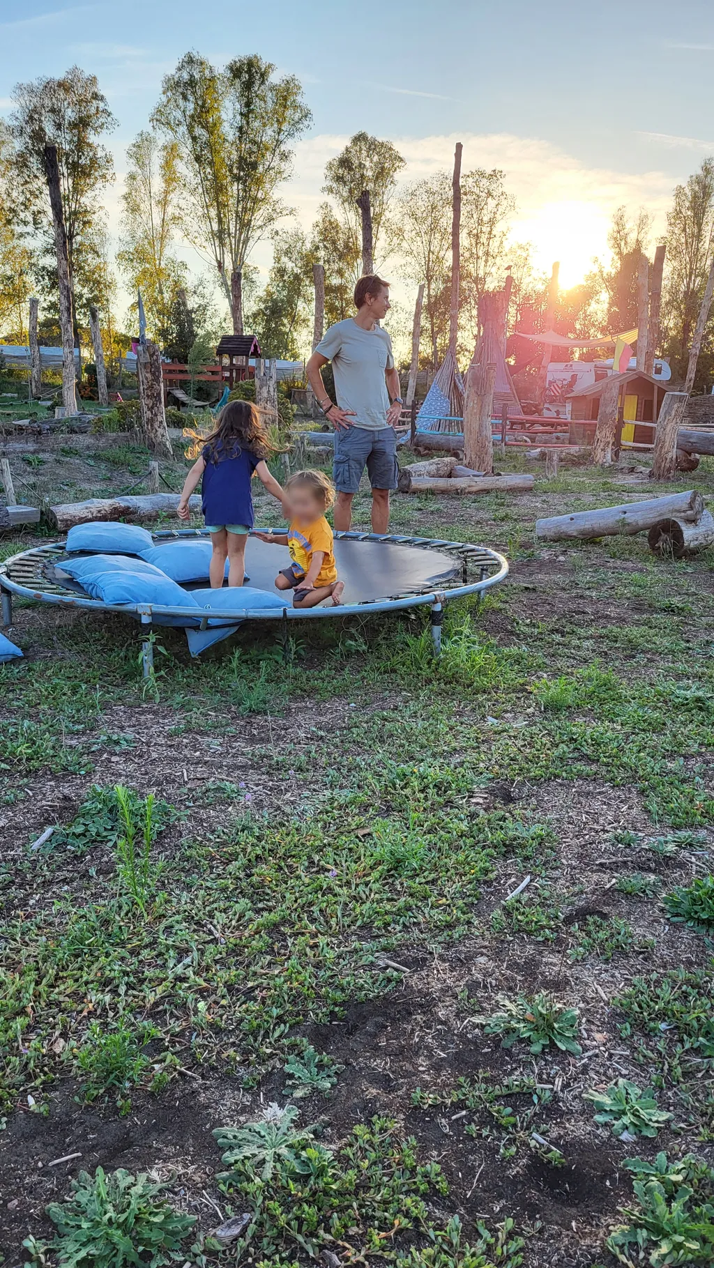 Un uomo e due bambini su un tappeto elastico controluce, nel sole della sera.