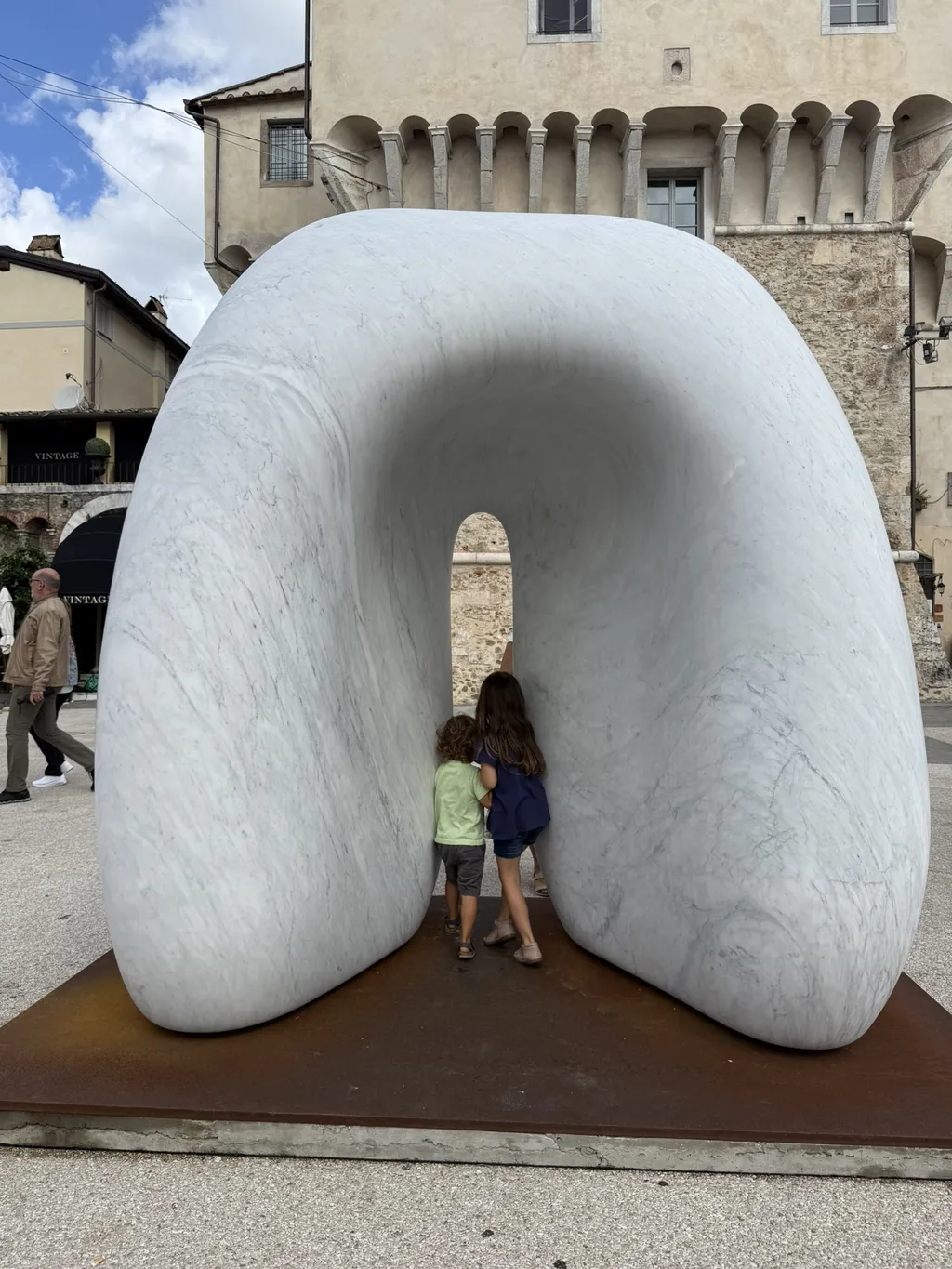 Un grande arco di marmo bianco in una piazza, dentro ci sono due bambini.