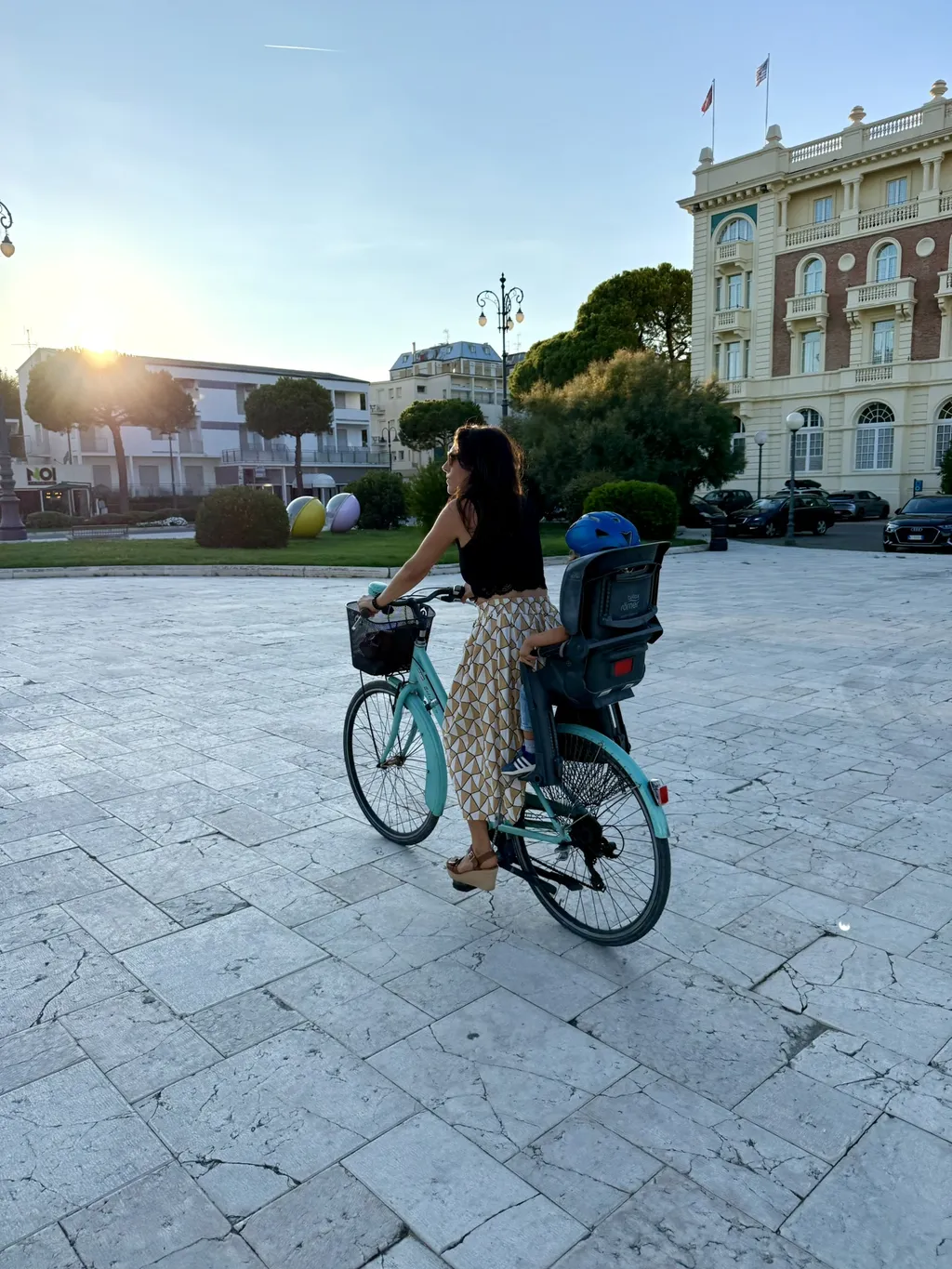 Una donna pedala con il seggiolino attraverso una piazza controluce.