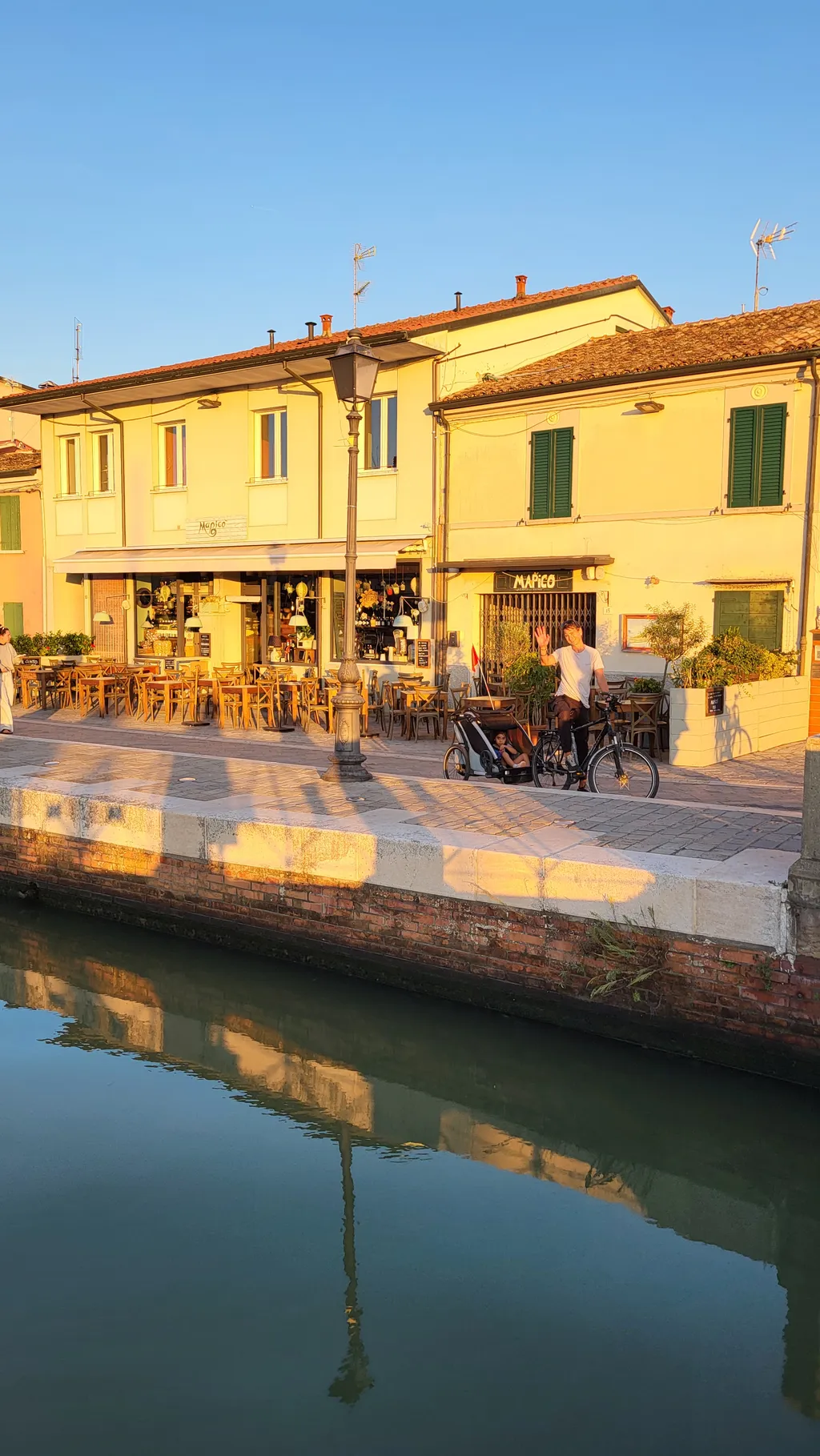 Un canale nella luce della sera, accanto una casa gialla con dei tavoli davanti.